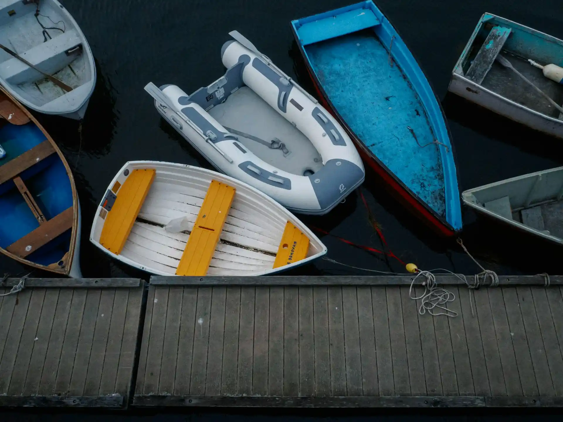 An overhead view of several small boats of various designs and colors, including wooden and inflatable, docked in dark water next to a wooden pier, illustrating a range of boat types throughout the evolution of boat paint.