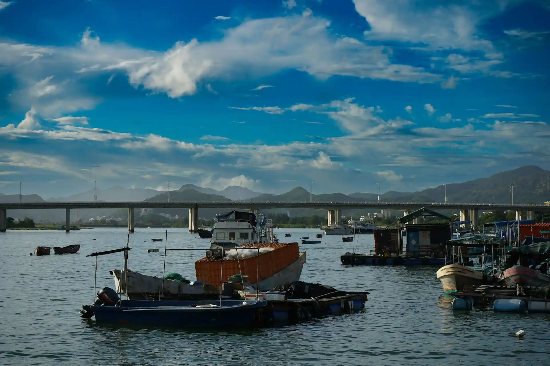 Various boats, including a large fishing vessel and a floating platform, anchored in a busy harbor with a bridge and mountains in the background, a setting that can influence do houseboats rock a lot.