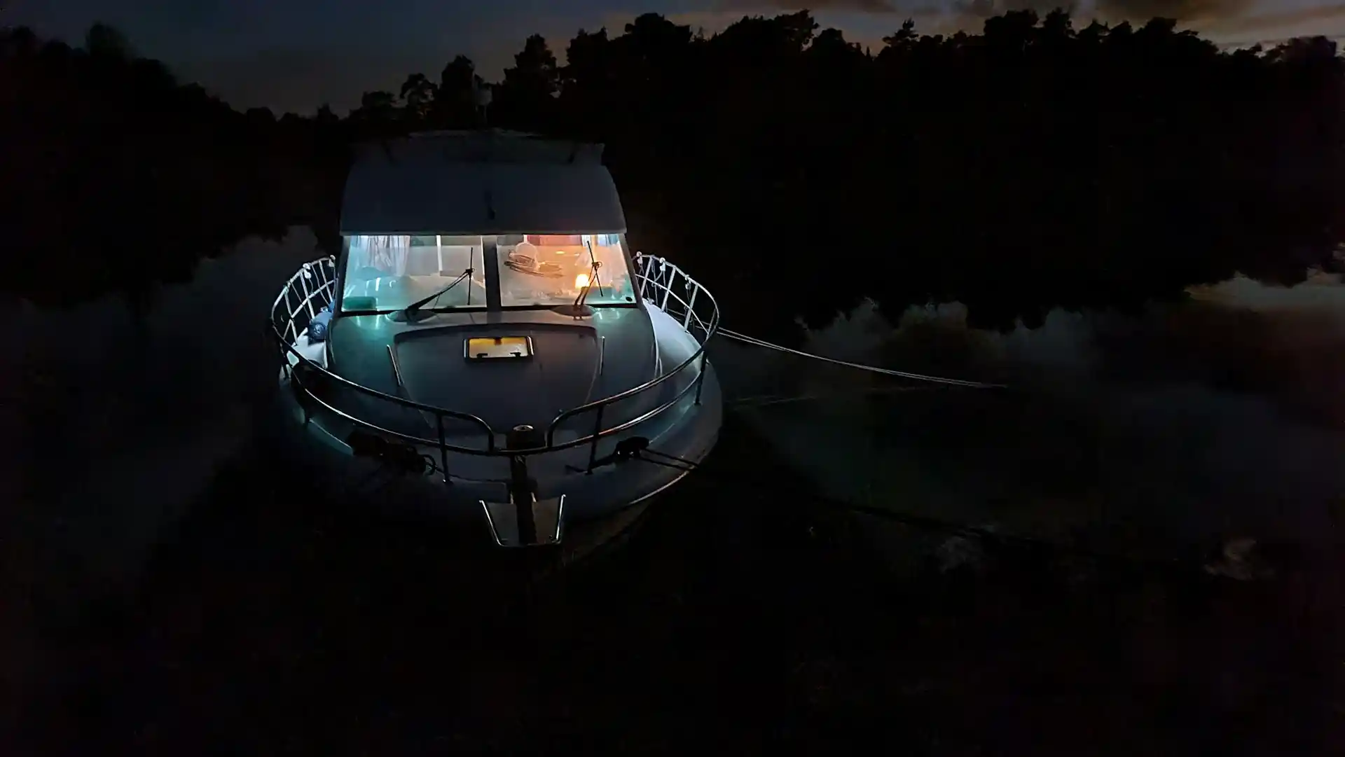 A houseboat illuminated by interior lights at night, docked in dark water with a treeline in the background, visually demonstrating how do houseboats get electricity.